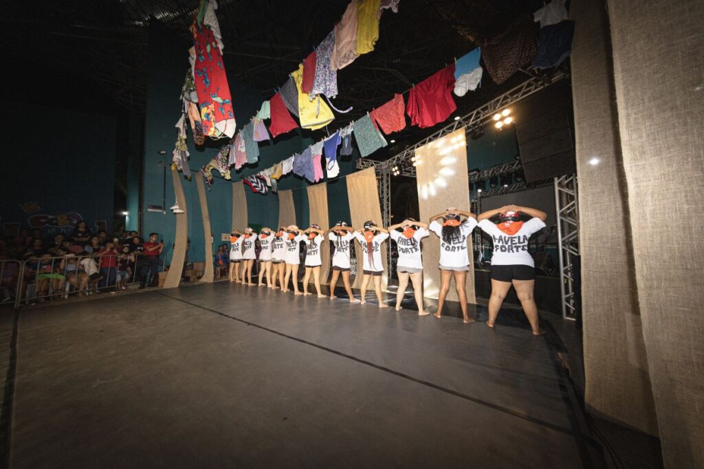 Na imagem, um grupo de pessoas está de costas para a câmera, alinhadas no palco e com as mãos sobre a cabeça, criando uma pose unificada. Elas usam camisetas com frases que não estão totalmente visíveis. Acima do palco, uma variedade de roupas coloridas está pendurada, como se fossem varais de lavanderia, contribuindo para uma atmosfera informal e festiva. Um público assiste atentamente à apresentação, com expressões de envolvimento. A iluminação do palco destaca os performers contra um ambiente mais escuro.