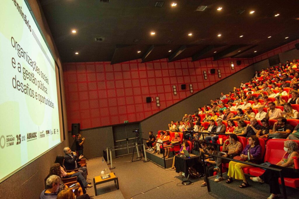 A foto mostra uma palestra acontecendo em um auditório com assentos vermelhos ocupados por espectadores. Uma pessoa está em pé ao lado de uma tela de projeção que exibe um slide com texto sobre organizações sociais e gestão pública. A iluminação suave e focada destaca o palestrante e a projeção. A audiência parece atenta ao conteúdo sendo apresentado. As paredes do auditório são de cor vermelha, e a atmosfera é de concentração acadêmica ou profissional.