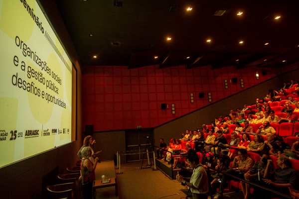 Nesta imagem, vemos uma palestra em um auditório com assentos vermelhos e paredes de cor correspondente. O público, variado e atento, preenche a maior parte dos assentos. No canto inferior esquerdo, o palestrante gesticula enquanto fala, dirigindo-se aos espectadores e a um grupo de pessoas sentadas à frente. Uma grande tela ao lado exibe um slide com texto relacionado a organizações sociais e gestão pública. A sala tem uma iluminação suave que foca na área do palestrante e da tela.