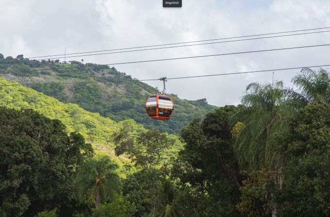 Um carro de teleférico paira no ar sobre um conjunto de elevações verdes iluminadas pelo sol