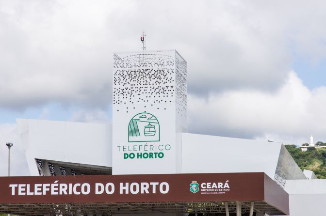 Uma torre branca está sobre uma base marrom. Em ambas está escrito Teleférico do Horto