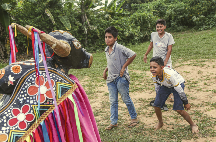 Fotografia com 3 crianças olhando para um boi cenográfico, típico da expressão cultural bumba-meu-boi. As crianças estão sorrindo.