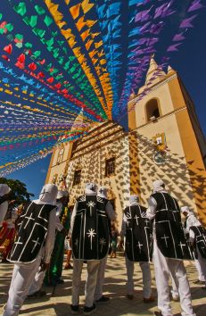 Uma igreja amarela enfeitada na rua com bandeiras coloridas e brincantes vestidos de preto e branco se aproximando do prédio.