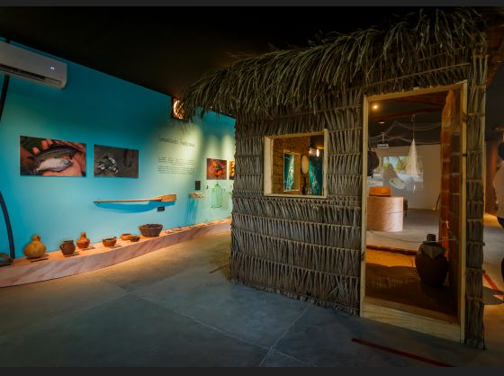 Uma casa de palha no meio de um museu de paredes azul-esverdeadas com artefatos de pesca e vida ribeirinha tradicional