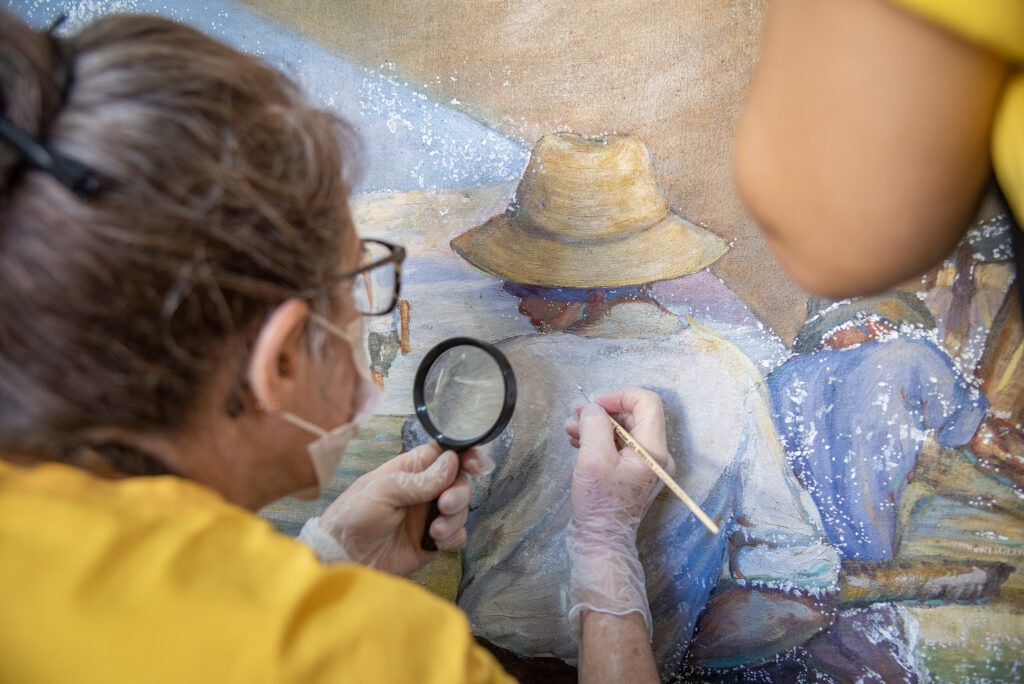 Imagem de uma mulher de óculos segurando uma lupa e restaurando uma pintura em tela. 