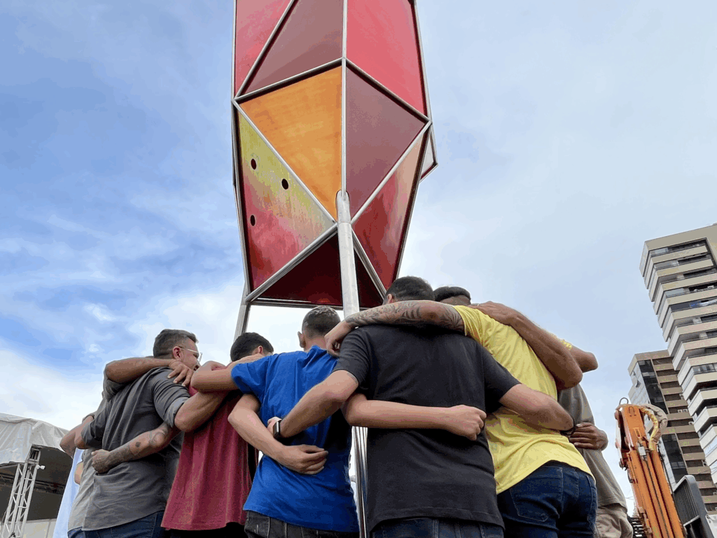 Grupo de pessoas de costas, em roda, abraçadas em gesto de união, debaixo de uma estrutura metálica vermelha ao ar livre. Ao fundo, céu azul e prédios.