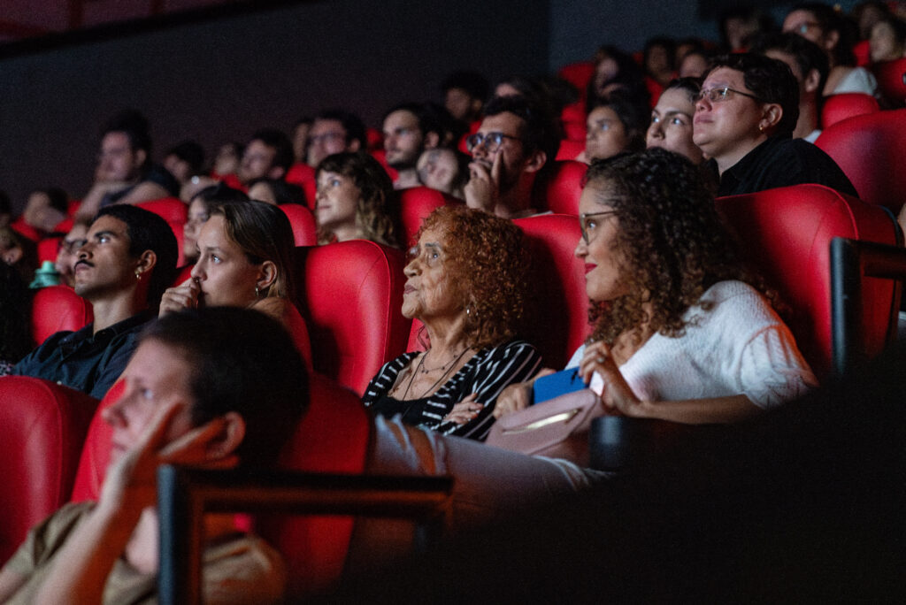 Público atento assiste a uma sessão de cinema em uma sala com poltronas vermelhas. As expressões variam entre concentração, curiosidade e emoção, refletindo o envolvimento das pessoas com o que está sendo exibido na tela.
