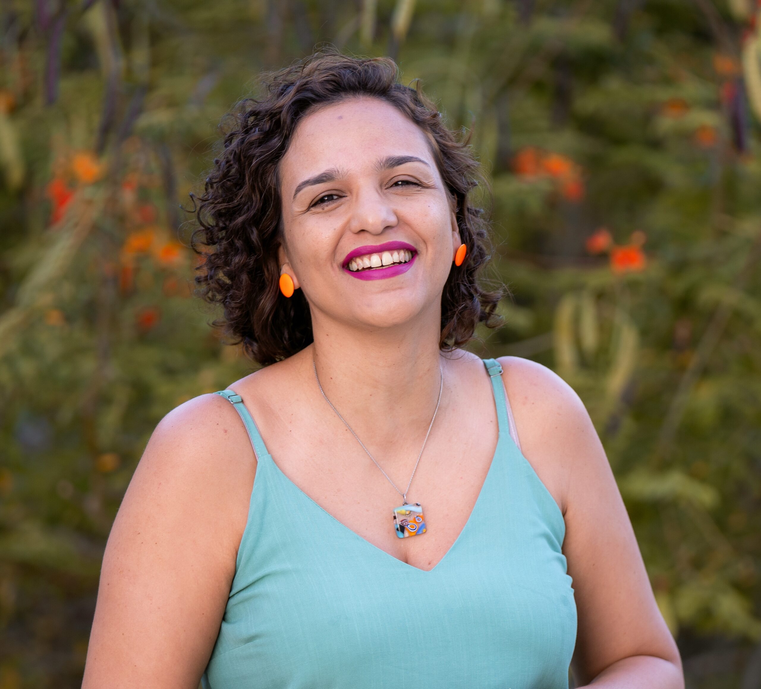 Mulher de cabelos curtos, cacheados e castanhos. Ela está sorrindo, usando batom rosa vibrante e brincos redondos de cor laranja. Veste uma blusa verde-clara de alças finas e um colar com pingente colorido.