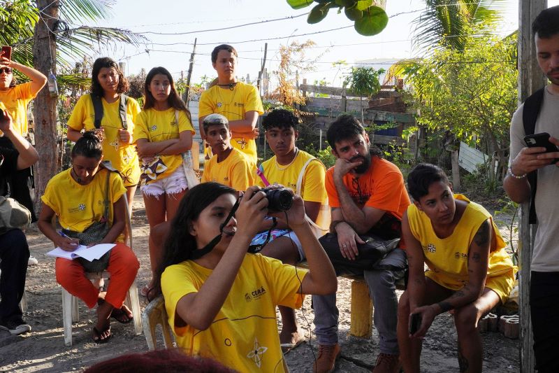 Grupo de jovens e adultos reunidos em espaço aberto, cercado por árvores e cercas de madeira. Vestindo camisas amarelas de projeto comunitário, alguns observam atentos, outros fazem anotações ou registram com câmera fotográfica, em uma atividade de aprendizado coletivo.