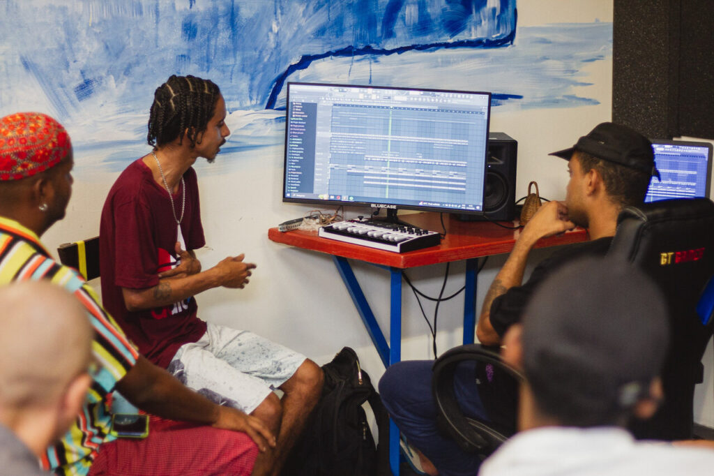 Grupo de jovens em uma sala de estúdio observa a tela de um computador com software de produção musical aberto. Um deles, sentado ao centro, explica algo enquanto gesticula, indicando o processo de criação de uma faixa.