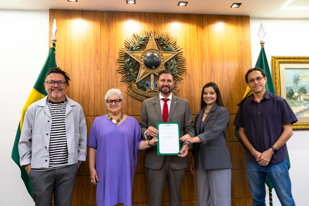Cinco pessoas posam sorrindo para a foto em frente ao brasão da República do Brasil. No centro, um homem de terno cinza e gravata vermelha segura um documento, acompanhado por uma mulher de blazer cinza. À esquerda e à direita, outras três pessoas completam o grupo, com bandeiras do Brasil ao fundo.