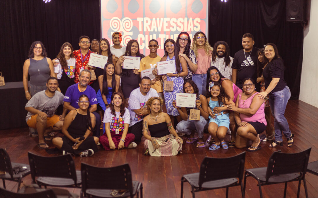 Grupo diverso de cerca de 25 pessoas posa sorridente em auditório, celebrando a conclusão do projeto Travessias Culturais. Algumas pessoas estão em pé, outras agachadas ou sentadas no chão, com certificados e pequenas sacolas nas mãos. Ao fundo, painel com a identidade visual em vermelho e branco escrito “Travessias Culturais”. O clima é de alegria e confraternização.