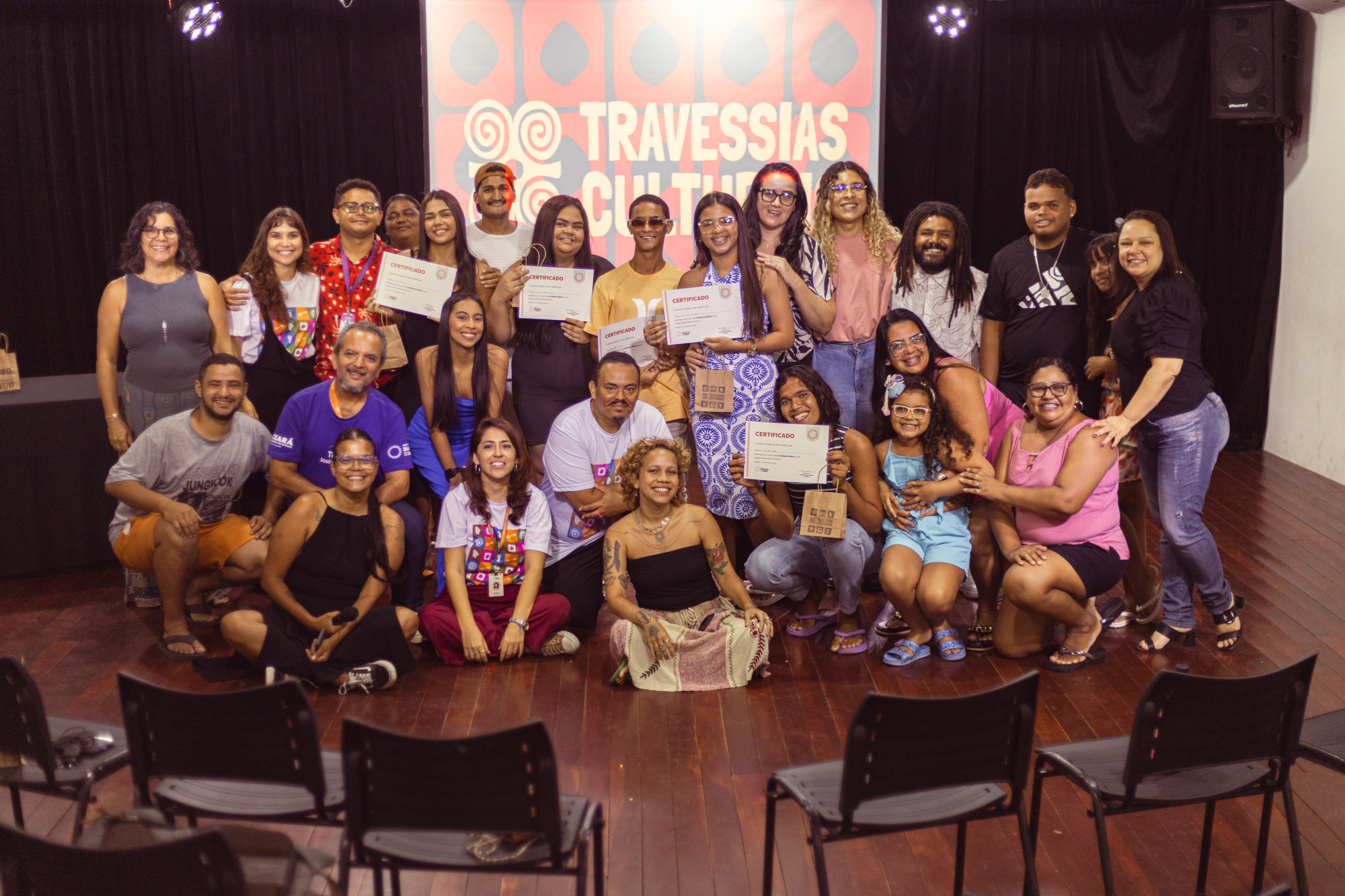 Grupo diverso de cerca de 25 pessoas posa sorridente em auditório, celebrando a conclusão do projeto Travessias Culturais. Algumas pessoas estão em pé, outras agachadas ou sentadas no chão, com certificados e pequenas sacolas nas mãos. Ao fundo, painel com a identidade visual em vermelho e branco escrito “Travessias Culturais”. O clima é de alegria e confraternização.