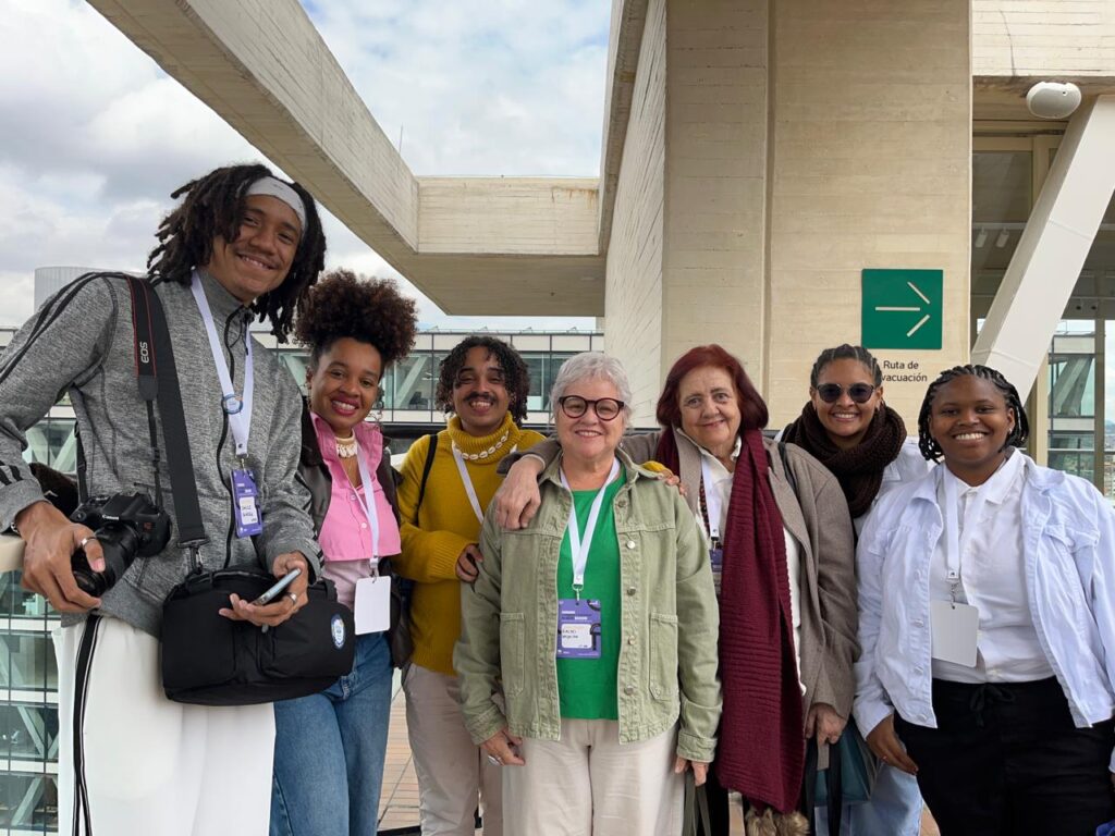 Um grupo de sete pessoas está posando sorridente para a foto em um ambiente externo, em frente a uma estrutura de concreto. Elas estão próximas umas das outras, transmitindo clima de descontração e amizade.