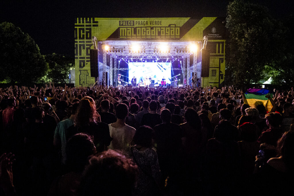 Grande público assiste a um show noturno no palco Praça Verde da Maloca Dragão, com iluminação intensa e músicos se apresentando no palco.