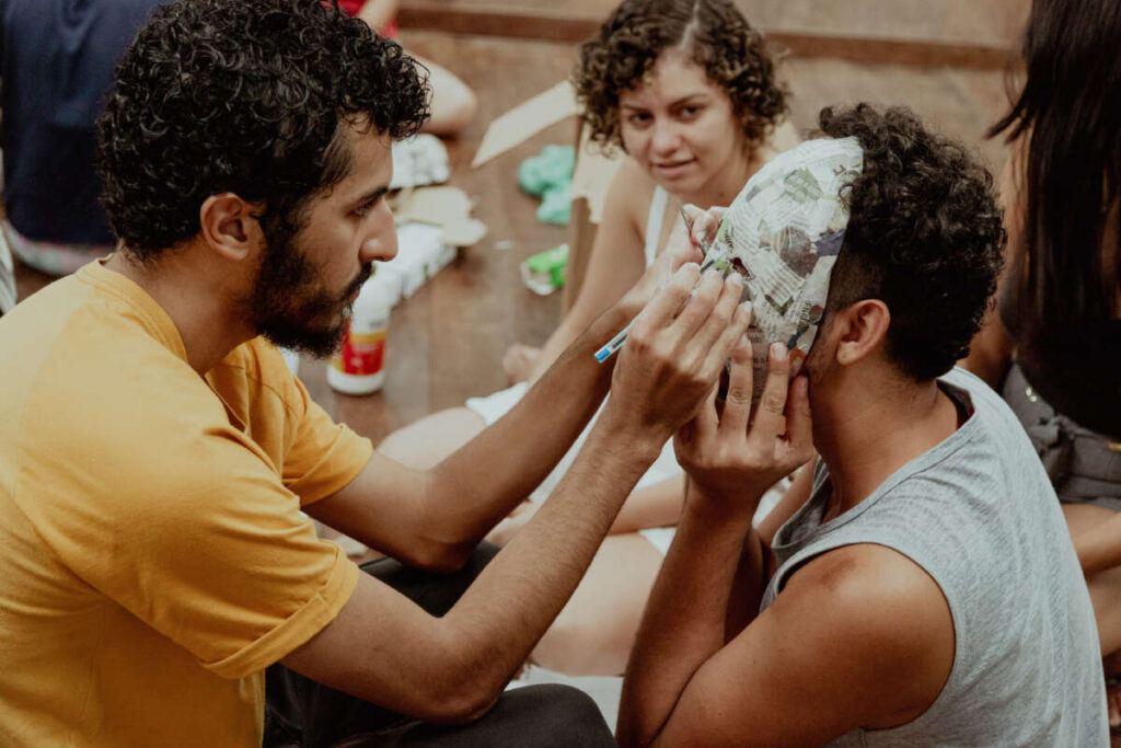 Pessoa com camiseta amarela pinta uma máscara de papel machê no rosto de outra pessoa sentada à sua frente. Ao fundo, outras pessoas observam a atividade.
