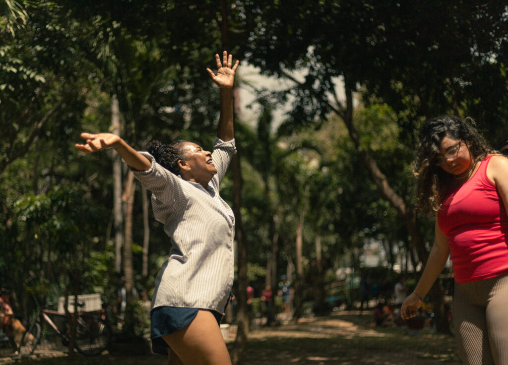A imagem mostra uma mulher pulando de alegria, com os braços levantados, no Parque do Cocó. Ela está cercada por árvores e natureza, transmitindo uma sensação de felicidade e liberdade ao ar livre.