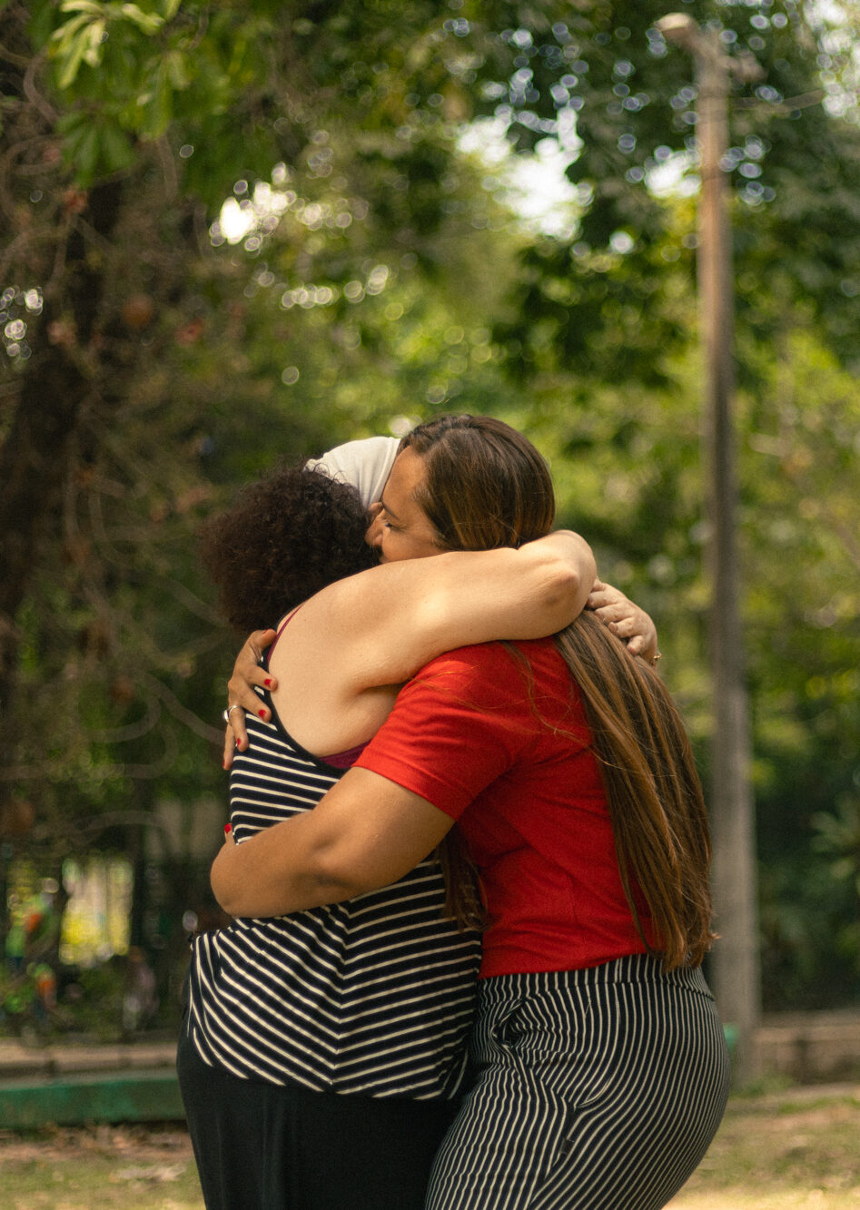 A imagem mostra duas mulheres se abraçando no Parque do Cocó, com árvores ao fundo. Uma mulher veste uma blusa de listras e um lenço na cabeça, enquanto a outra usa uma camisa vermelha. A cena transmite uma sensação de carinho e amizade.