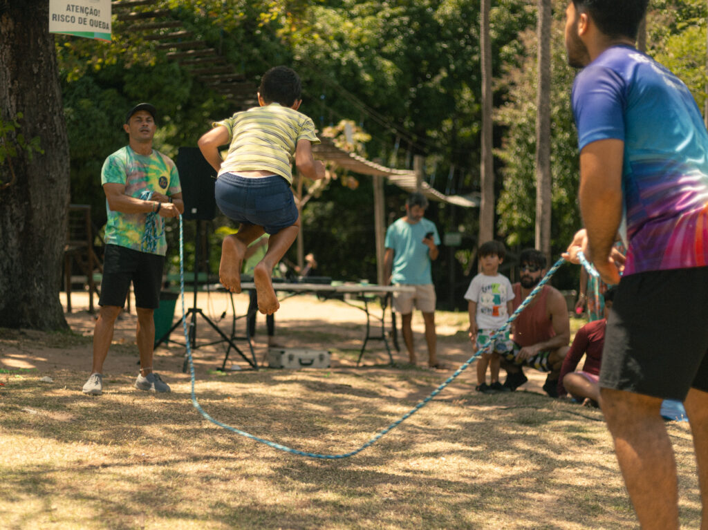 A imagem mostra duas pessoas girando uma corda de pular ao ar livre, no Parque do Cocó. Uma delas está no centro, saltando com entusiasmo, enquanto a outra ajuda a girar a corda. Ao fundo, várias pessoas observam e participam da atividade, cercadas por árvores e vegetação