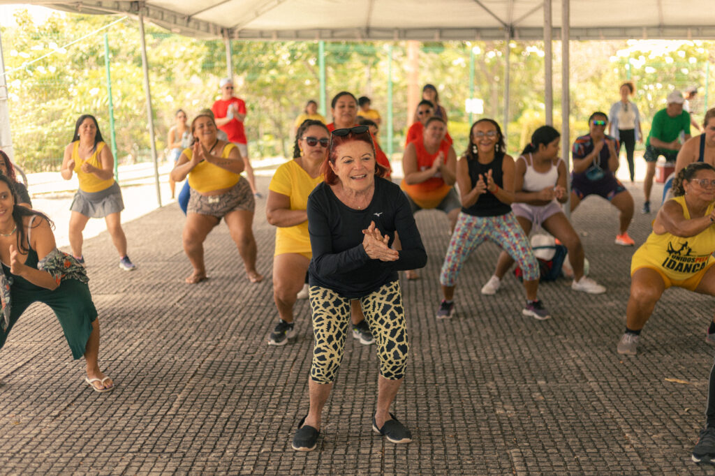 Um grupo de pessoas de várias idades participando de uma aula de dança ou atividade fitness ao ar livre, sob uma estrutura coberta. As pessoas estão sorrindo, se movimentando e se divertindo. Ao fundo, há árvores e um ambiente natural do Parque do Cocó.