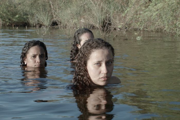 Três mulheres estão parcialmente submersas em um rio, com a água na altura dos ombros. A que está mais à frente olha diretamente para a câmera, com expressão séria. As outras duas aparecem atrás dela, também com semblantes atentos. O ambiente é natural, com vegetação rasteira e margens cobertas por capim. A luz suave ressalta o tom sereno e misterioso da cena.