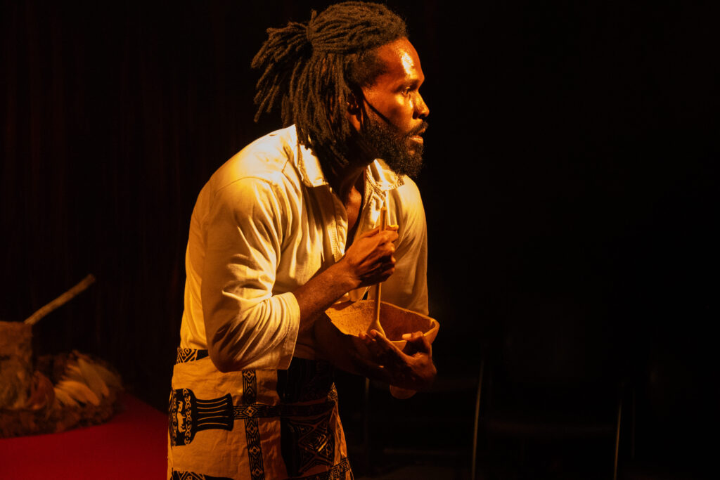 Pessoa em cena teatral, com expressão concentrada, segura uma tigela de barro e um bastão de madeira. Ela usa camisa clara e tecido estampado amarrado na cintura. A iluminação amarelada destaca o rosto e o figurino, criando um clima intenso e intimista no palco escuro.