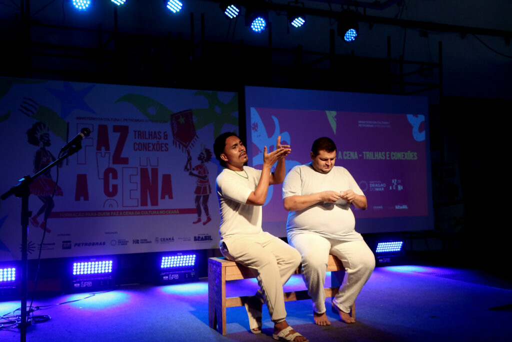 Dois homens estão sentados em bancos de madeira sobre um palco iluminado por luzes azuis e roxas. Ambos vestem roupas claras, em tons de branco e bege, e estão descalços, compondo uma cena de estética minimalista. O homem à esquerda gesticula com as mãos erguidas, como se estivesse explicando ou encenando algo, com expressão concentrada e olhar voltado para frente. O homem à direita mantém a cabeça levemente abaixada, focado em um objeto pequeno que segura nas mãos. Ao fundo, há painéis com a identidade visual do evento “Faz a Cena – Trilhas e Conexões”, com logotipos institucionais e grafismos coloridos, indicando um contexto de apresentação cênica ou formação cultural em um espaço teatral.