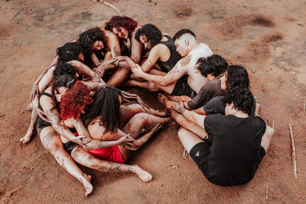 Grupo de pessoas sentadas no chão em círculo, com os corpos próximos e entrelaçados, apoiando-se mutuamente. Algumas estão com o corpo coberto de terra, sugerindo uma ação coletiva ou performance. A cena transmite conexão, cuidado e união entre os participantes.
