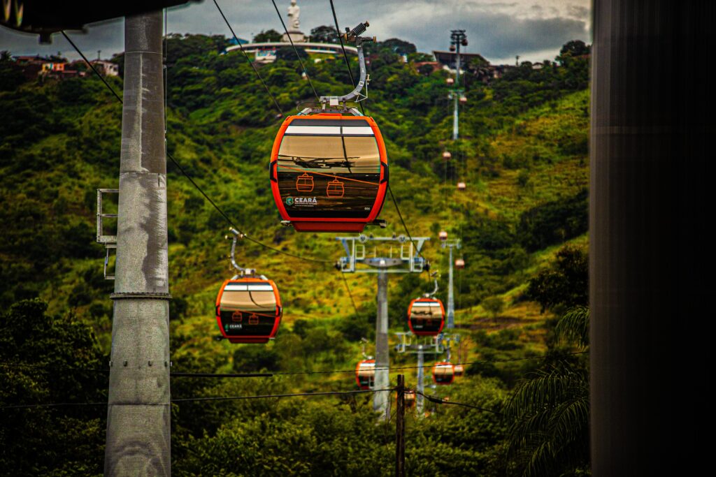 Cabines de teleférico nas cores laranja e preto atravessam uma paisagem verde de serra, suspensas por cabos entre torres metálicas. Ao fundo, colinas cobertas de vegetação e construções se espalham pelo horizonte sob um céu nublado.

