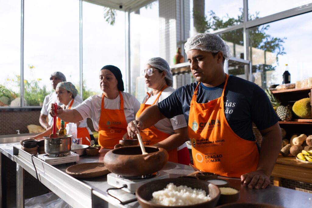 Grupo de pessoas em cozinha iluminada prepara alimentos de forma colaborativa, usando toucas e aventais laranja. Em primeiro plano, um participante mexe uma panela de barro, enquanto outros cuidam de diferentes etapas da receita, com ingredientes e utensílios organizados na bancada.
