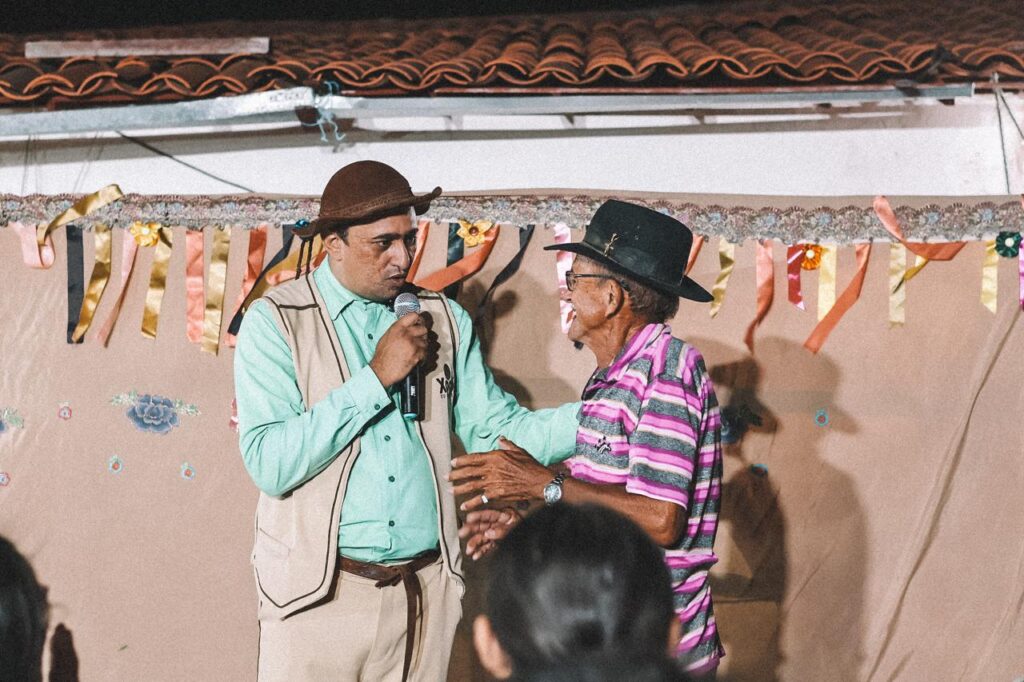 Homem vestido com traje típico nordestino, usando chapéu e segurando um microfone, conversa sorrindo com um idoso de chapéu e camisa listrada em um palco decorado com bandeirinhas coloridas. Ao fundo, há uma parede simples e o telhado de uma casa, sugerindo um ambiente de festa popular. Algumas pessoas assistem à cena em primeiro plano.