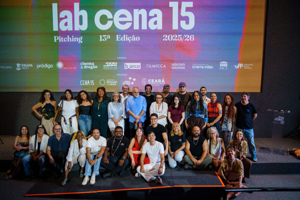 Foto de grupo com cerca de 30 pessoas no palco de um auditório, posando sorridentes após uma apresentação. Ao fundo, um telão colorido exibe o texto “lab cena 15 – Pitching – 13ª edição 2025/26”, junto a logotipos de instituições parceiras. O grupo está organizado em duas fileiras, com parte das pessoas em pé e outras sentadas ou agachadas na frente. À direita, há um púlpito com microfone, indicando o contexto de evento ou apresentação.
