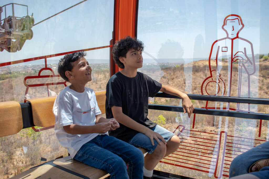 Dois meninos sentados dentro de um teleférico observam a paisagem seca ao redor, com vegetação esparsa e vista ao fundo. Eles parecem atentos e curiosos. No vidro da cabine há ilustrações em vermelho que representam personagens e elementos do equipamento.
