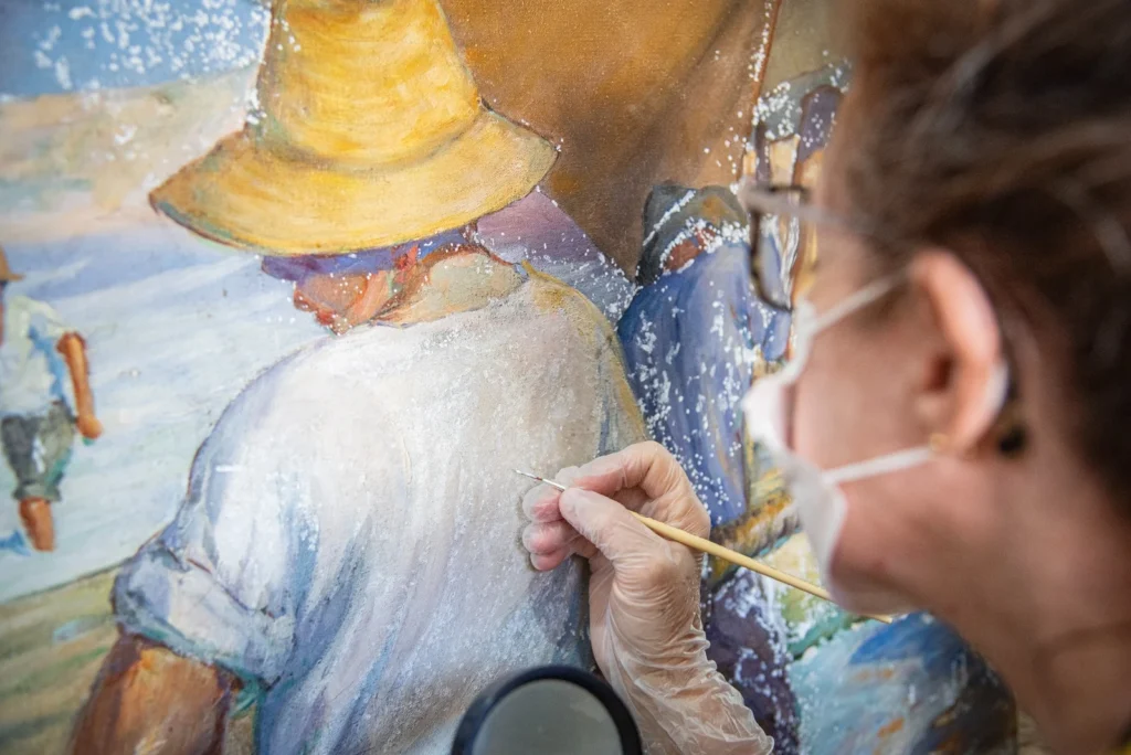 Fotografia em plano detalhe e ângulo lateral de uma mulher realizando o trabalho de restauração em uma pintura em tela. À direita, em primeiro plano e levemente desfocada, está a restauradora de perfil. Ela usa óculos de grau, máscara de proteção branca e luvas descartáveis transparentes. Com a mão direita, ela segura um pincel fino de cabo longo, tocando delicadamente as costas de um personagem pintado na tela. A pintura retrata trabalhadores, possivelmente pescadores, vestindo roupas claras e chapéus de palha. O fundo da obra traz tons de azul e amarelo.