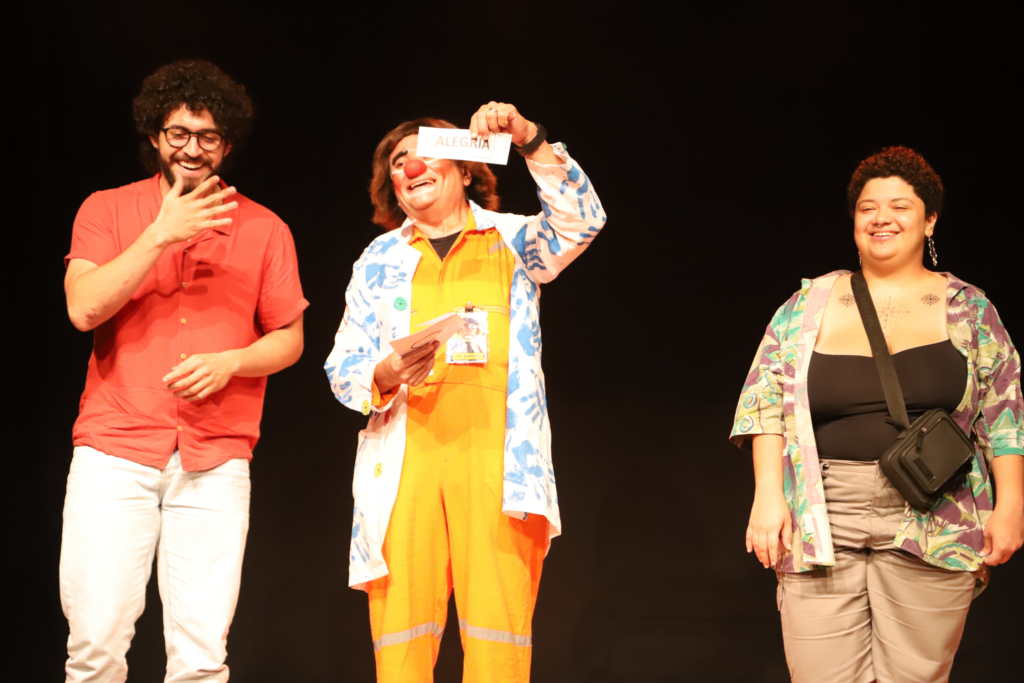 Três pessoas sorriem em um palco com fundo preto. À esquerda, um homem de óculos e camisa vermelha ri com a mão perto do rosto. No centro, uma pessoa vestida de palhaço, com macacão laranja, jaleco decorado e nariz vermelho, segura um papel acima da cabeça escrito "ALEGRIA". À direita, uma mulher de cabelos curtos, blusa estampada aberta e bolsa transversal sorri para a cena. A iluminação é focada no trio.